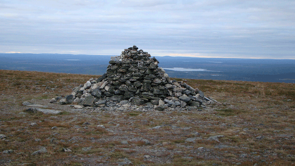Monument to Finnish Sisu. Rocks are arranged into a large mound at the edge of a grassy plateau, overlooking fields, forests, and lakes, under a bright cloudy sky.